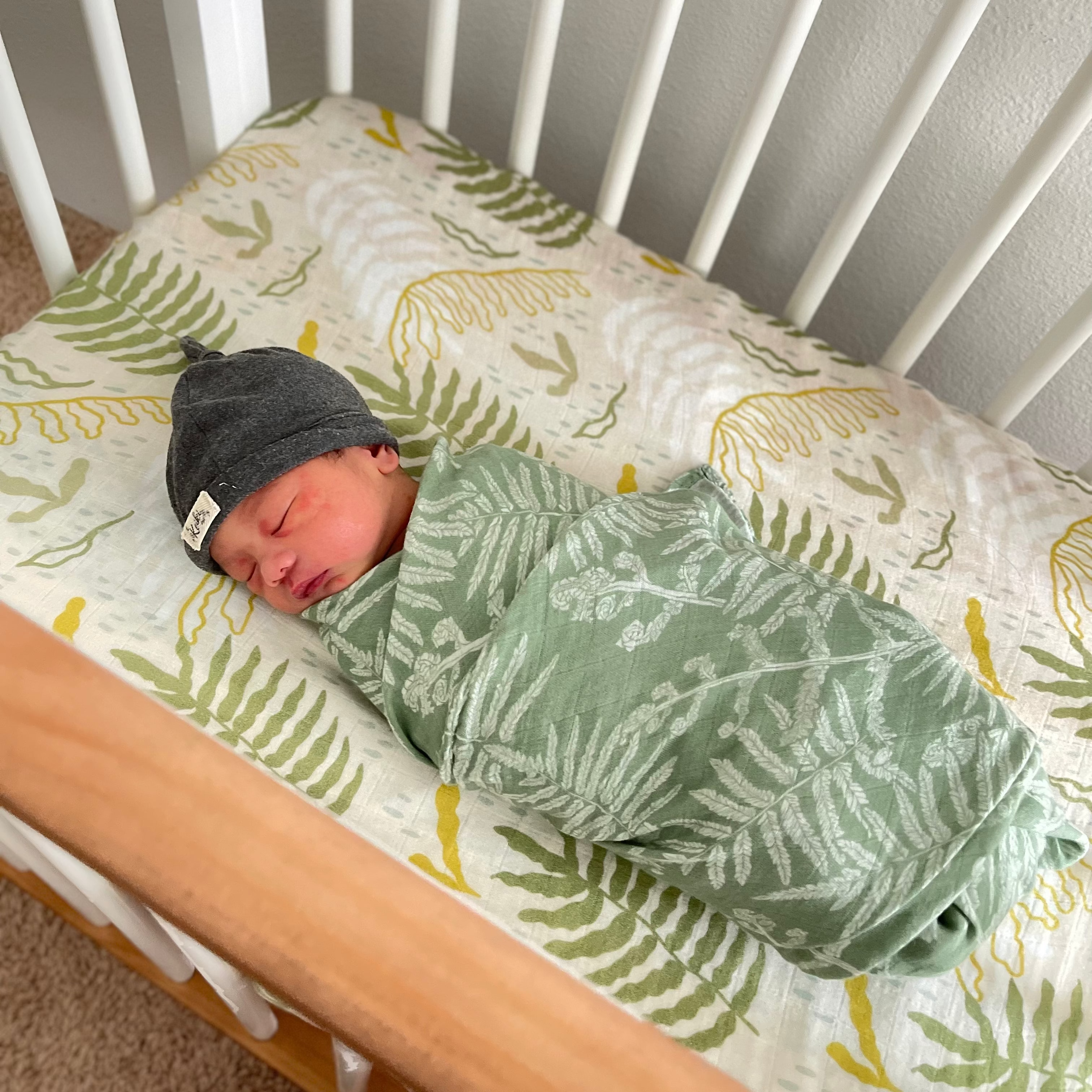 Newborn baby swaddled in a green blanket with fern pattern, sleeping in a crib with white bedding.