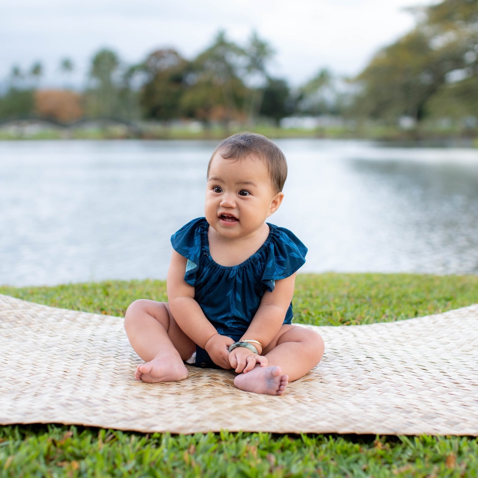 Baby sitting on a woven mat by the water with trees in the background