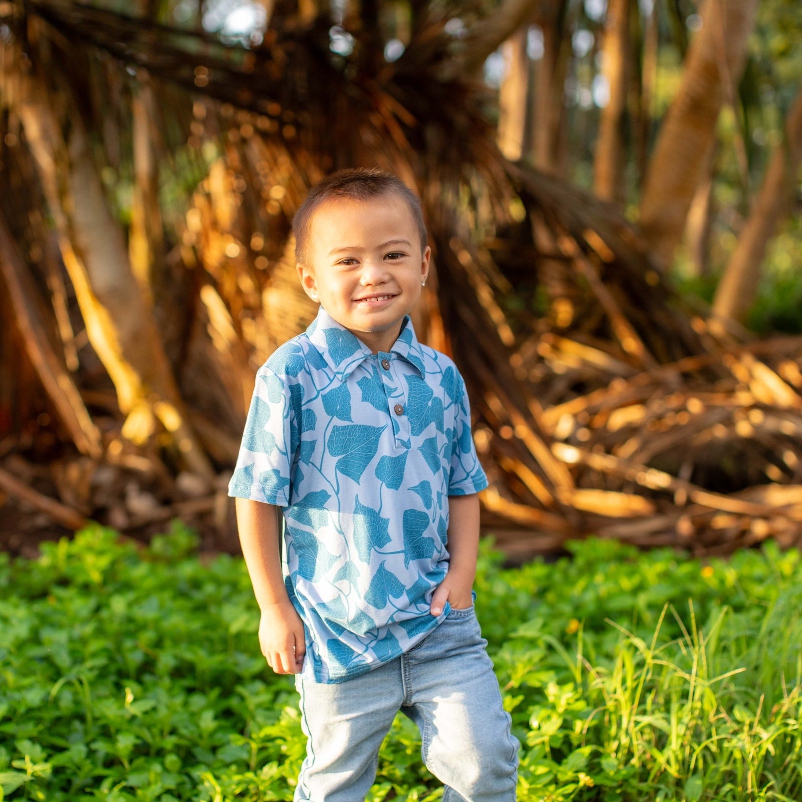 Child in a blue patterned shirt standing in a grassy area with trees in the background