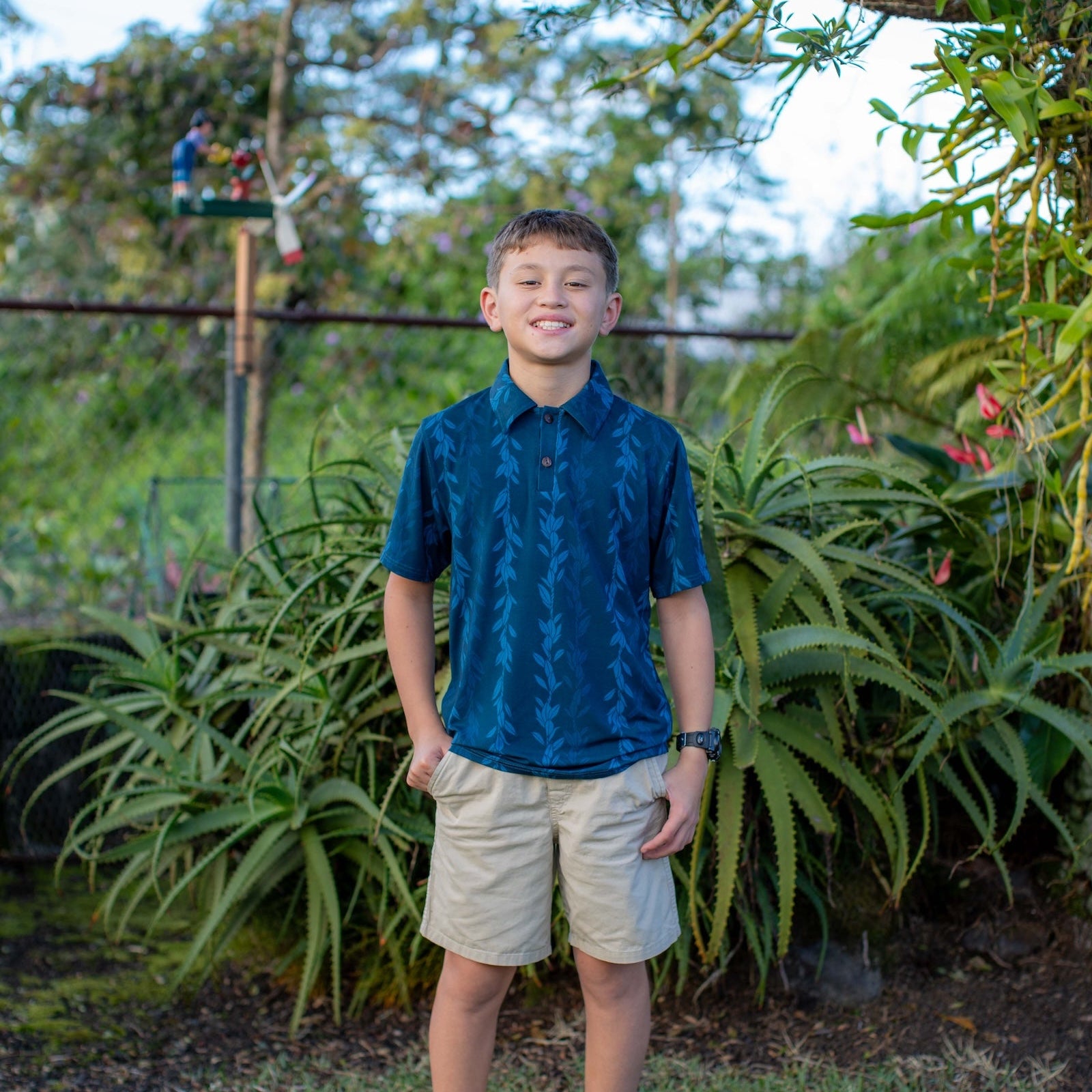 Young boy standing outdoors in a garden with greenery and plants around him.
