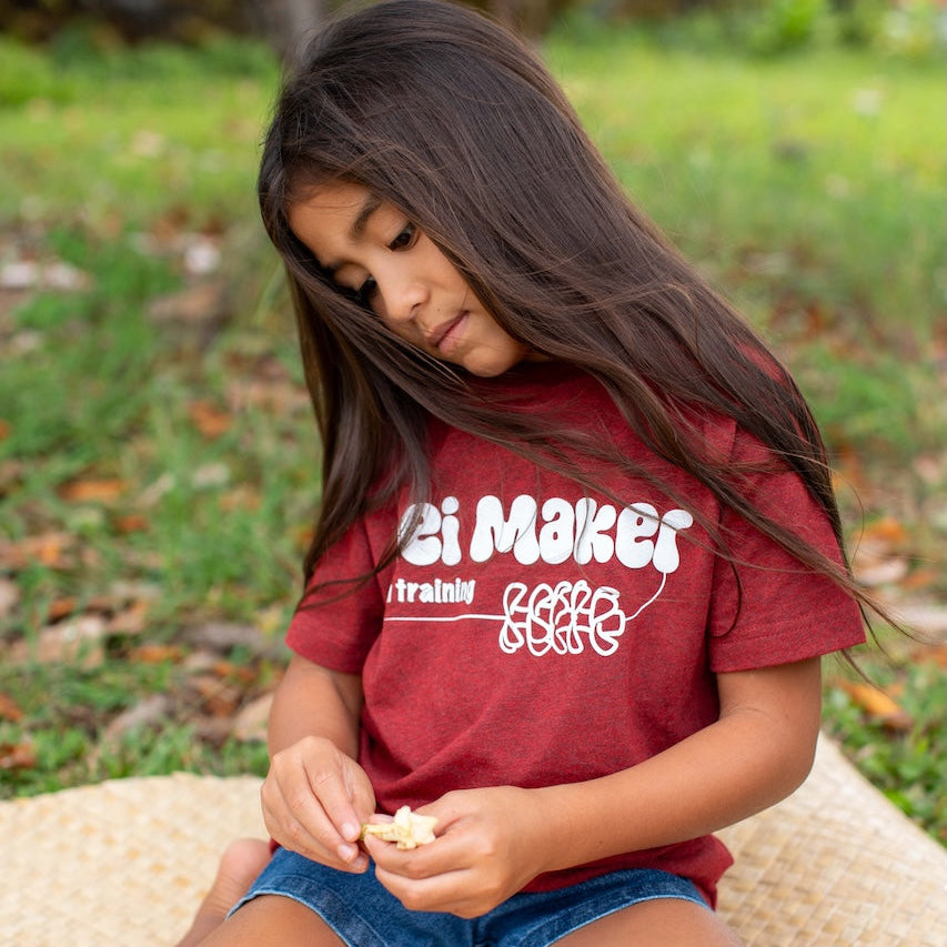 Young girl sitting outdoors wearing a red t-shirt with text making lei
