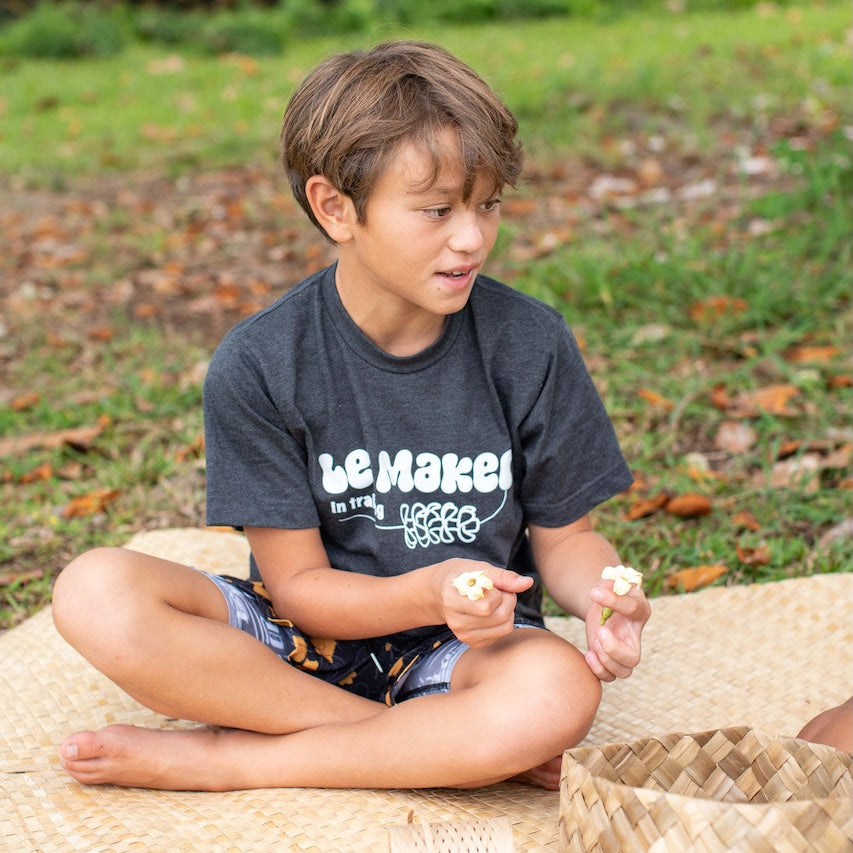 Child sitting on a woven mat outdoors, making lei