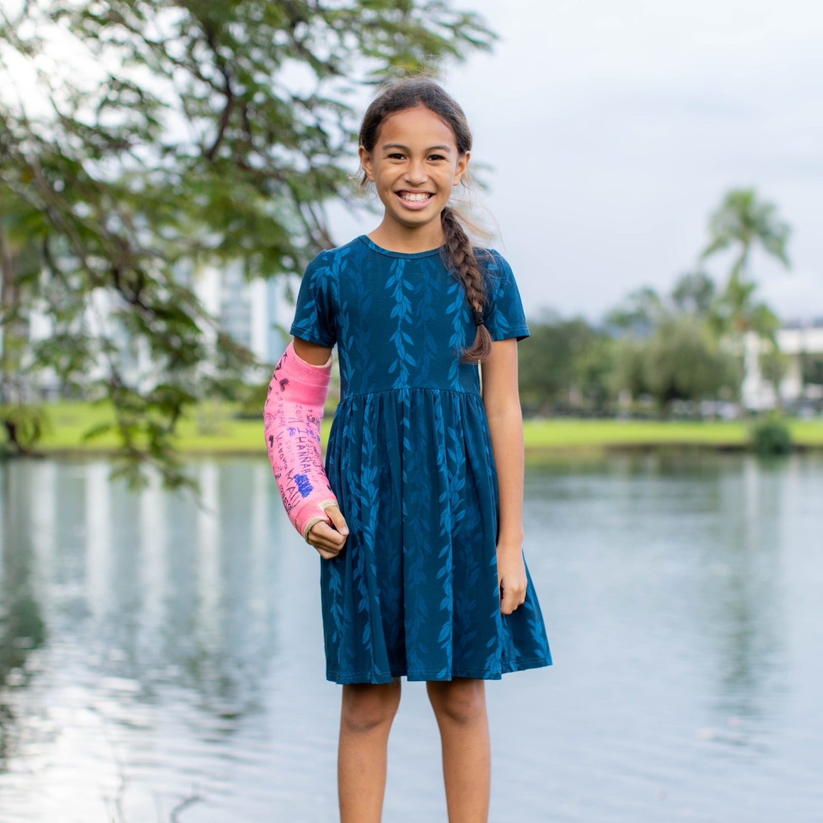 Young girl in a blue dress standing by a lake with a pink cast on her arm.