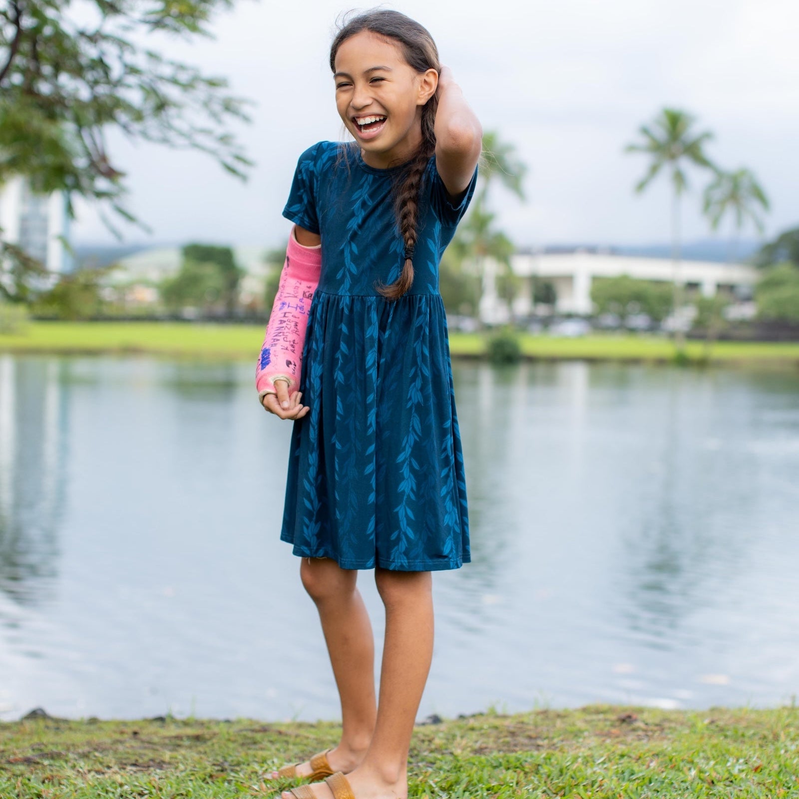 Girl in a blue maile strands dress standing by the water with trees in the background