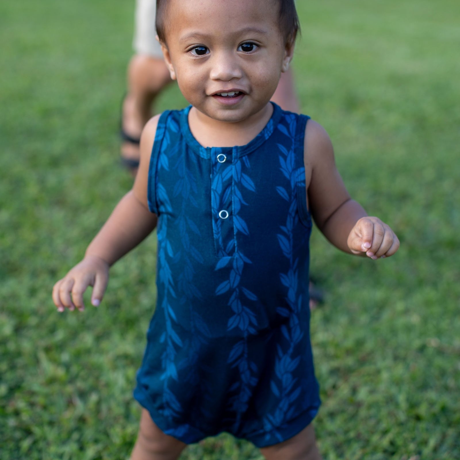 Child in a blue maile strands romper standing on grass with a boy in the background