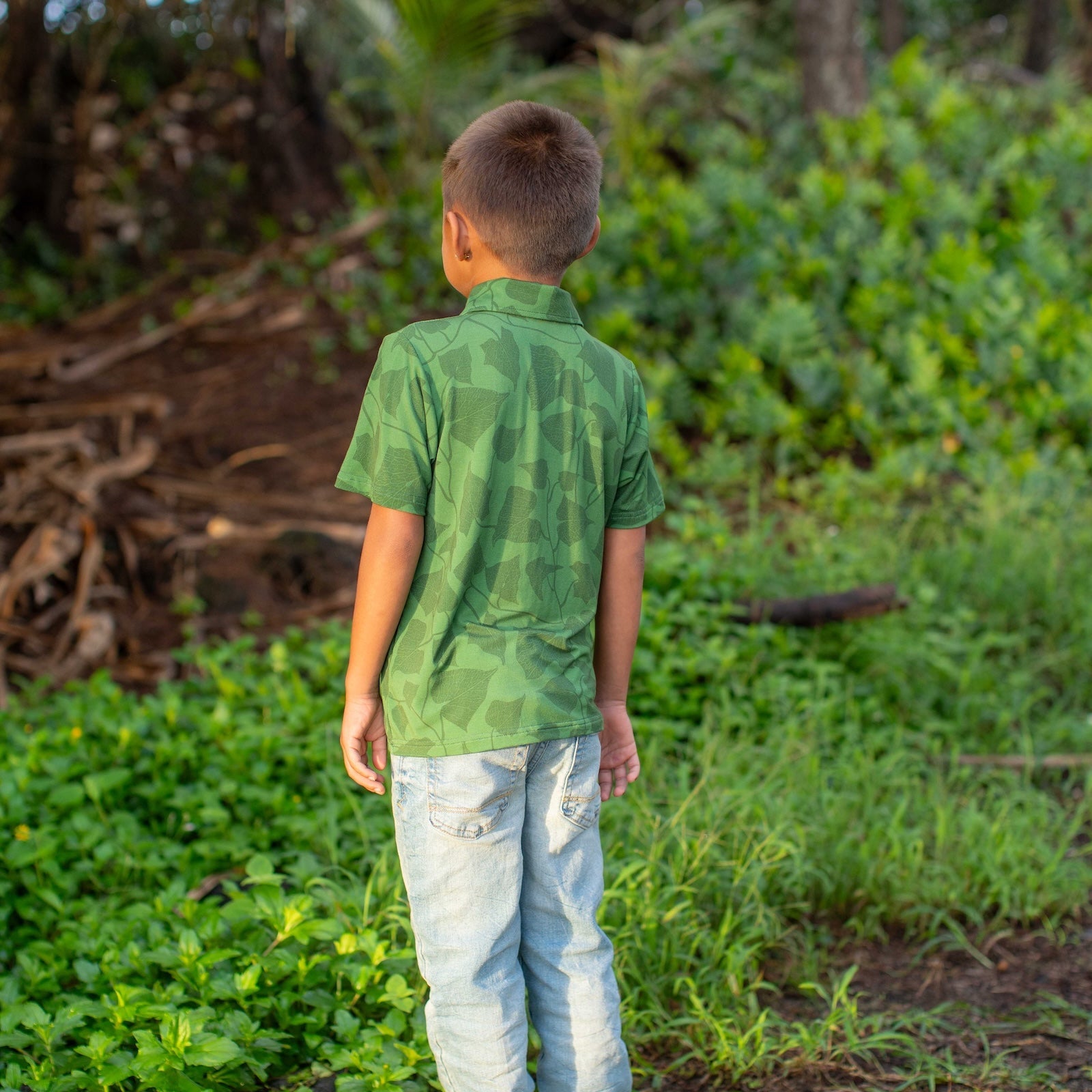 Child in a green peahi fern polo and blue jeans standing in a natural setting with greenery.