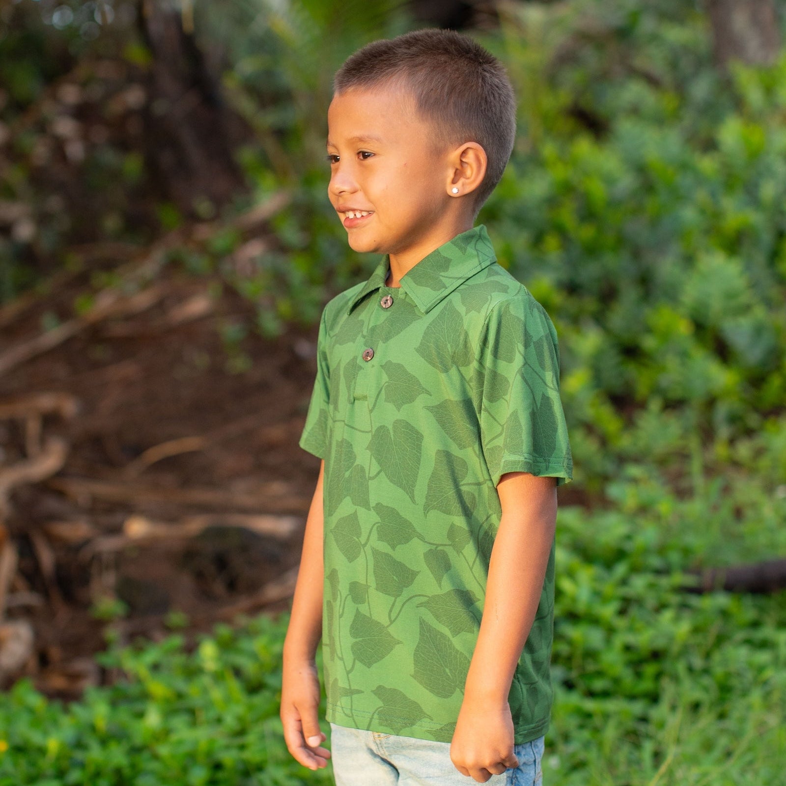 Young boy wearing a green peahi fern polo shirt standing in a natural setting with trees and foliage.