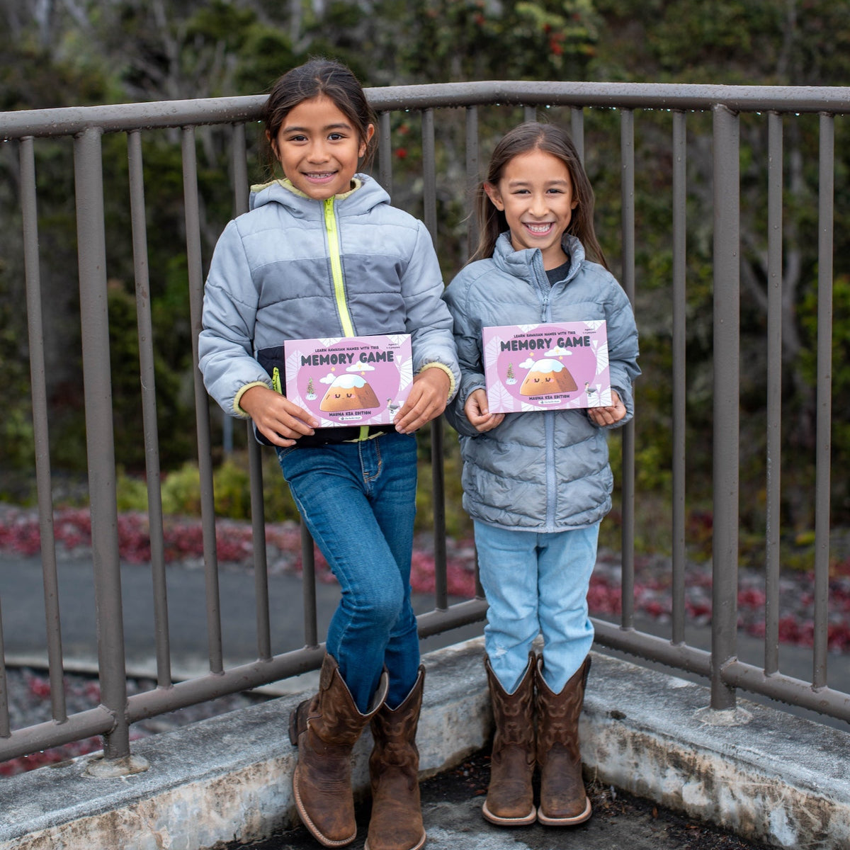 Two children holding pink game boxes with a metal railing and trees in the background.