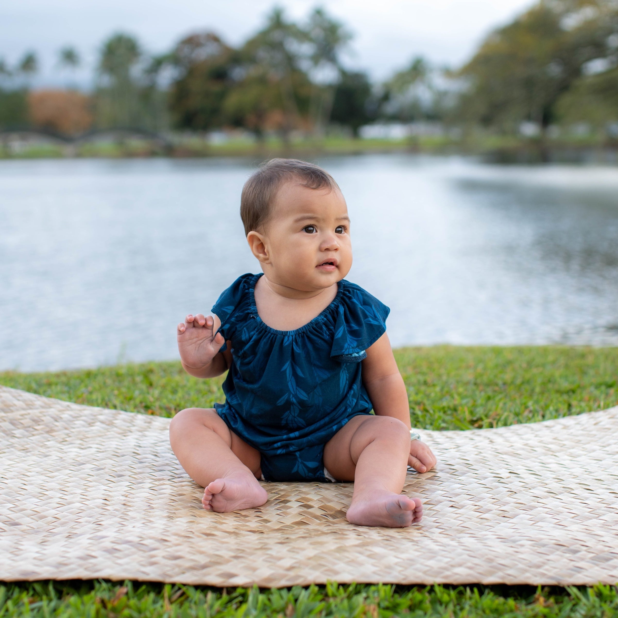 Baby in a blue maile strands flutter romper sitting on a woven mat by the water