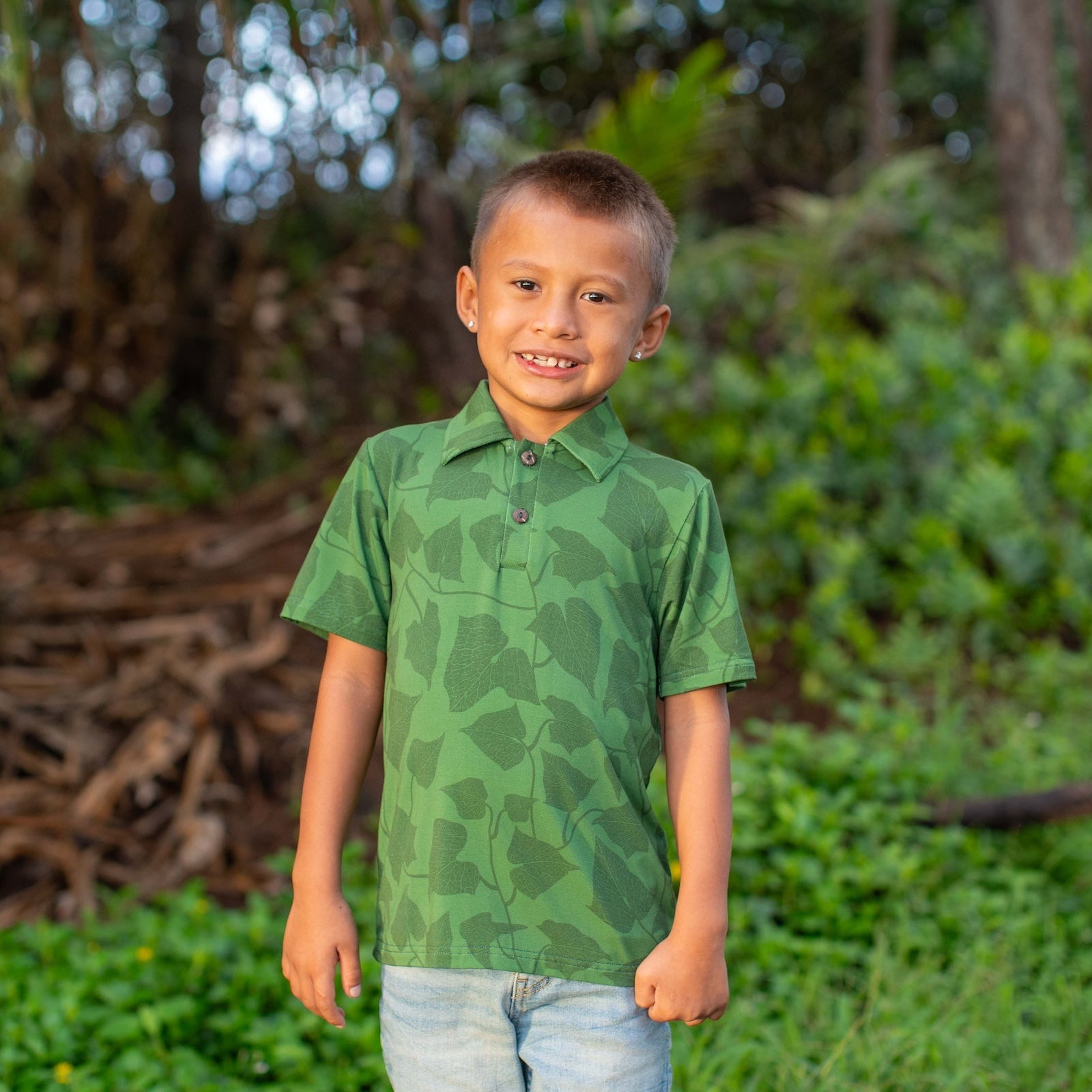 Young boy in a green peahi fern polo standing in a natural setting with trees and foliage.