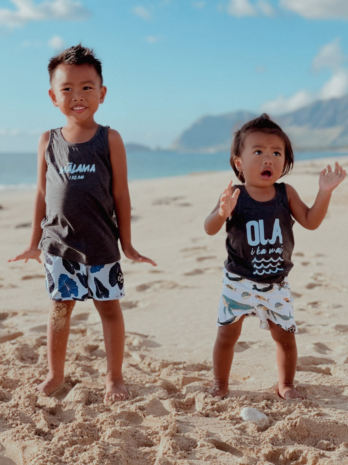 two brothers wearing their malama i ke kai and ola i ka wai tank tops. They are standing on the shoreline at the beach.