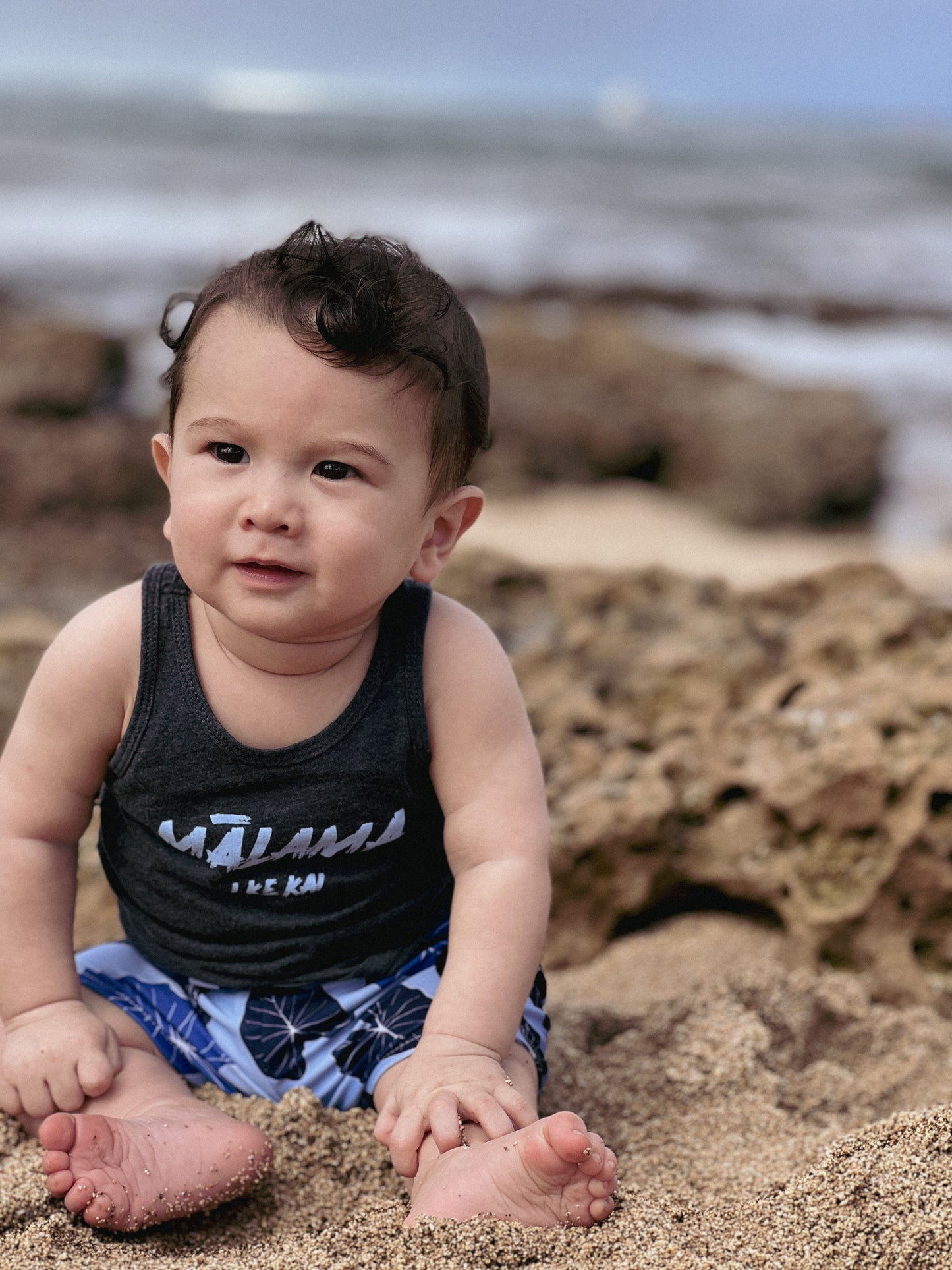 baby boy wearing a malama i ke kai tank top sitting on the beach. he is wearing blue kalo boardshorts.